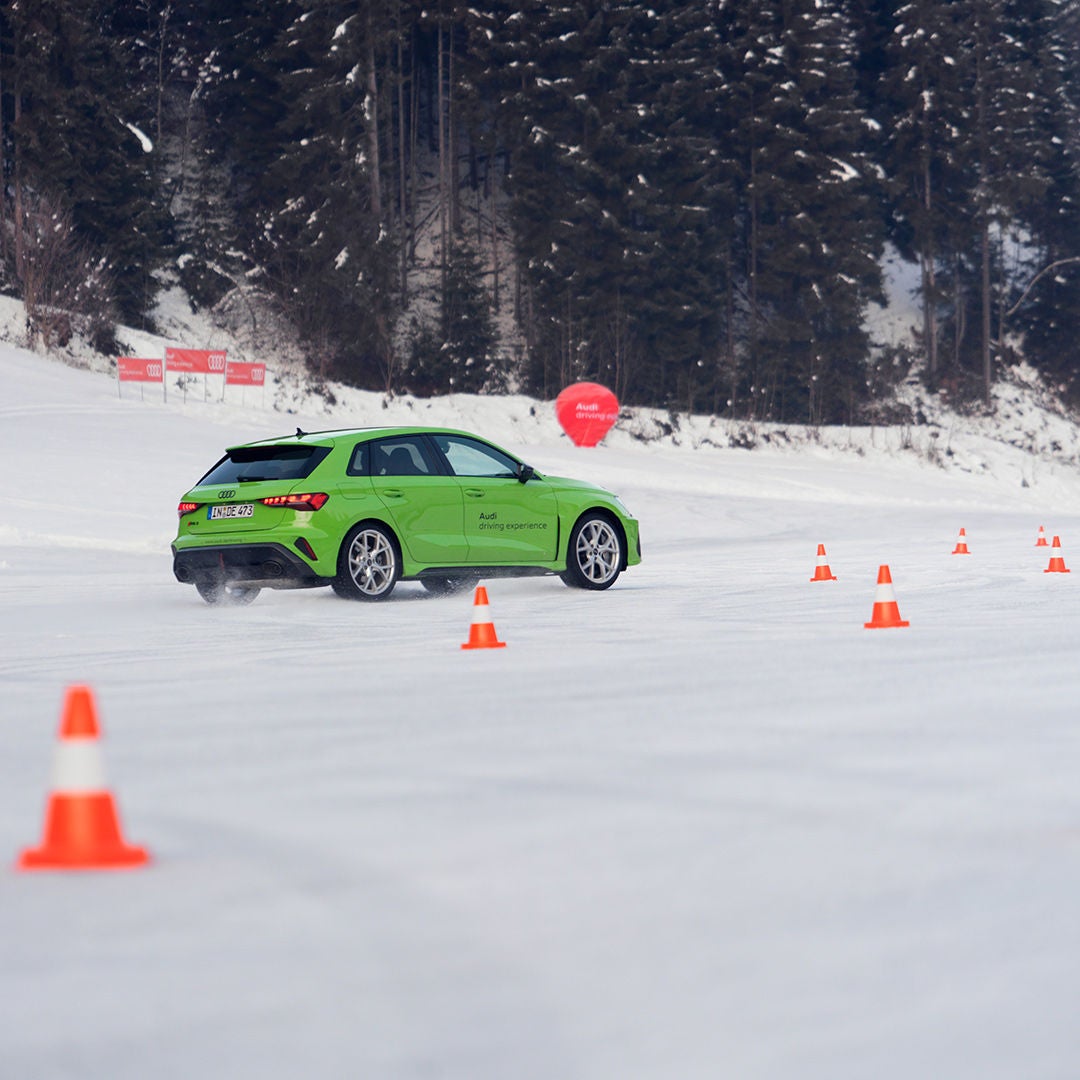 A light green Audi RS 3 Sportback drives along a snow-covered road between orange cones, with a snow-covered forest in the background.