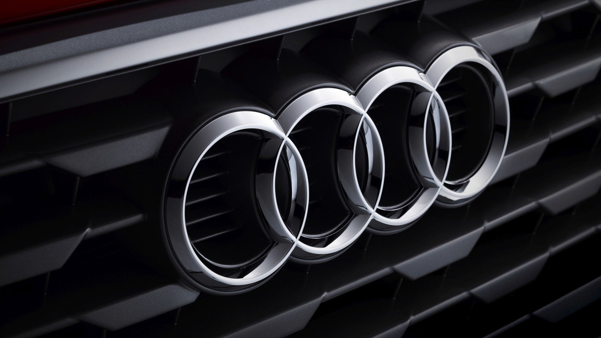 Nahaufnahme des Audi-Logos mit vier Ringen auf schwarzem Kühlergrill eines Fahrzeugs.          