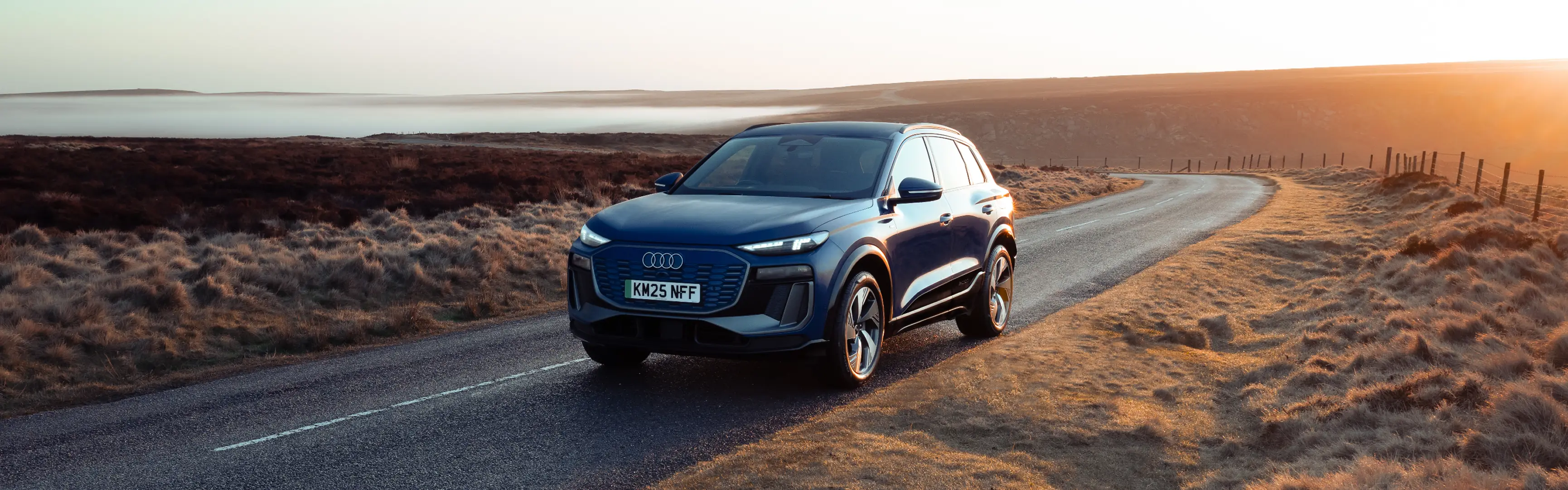 Blue Audi Q6 e-tron driving along a winding rural road at sunrise. Shown from the front three-quarter angle, the SUV features LED headlights, a wide front grille and aerodynamic alloy wheels. Golden morning light illuminates the surrounding moorland landscape, with mist and rolling hills visible in the distance as the road curves towards the horizon.