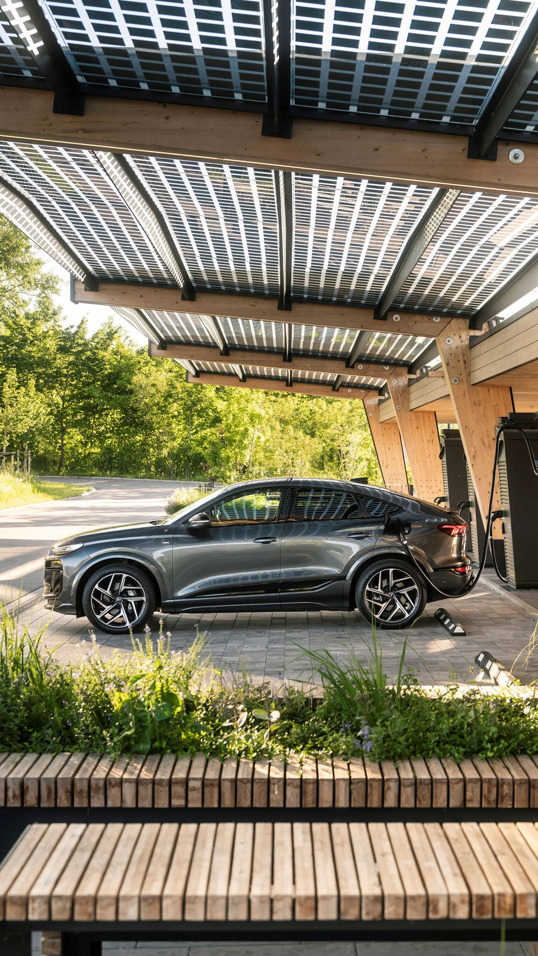 Dark grey Audi Q6 Sportback e-tron charging at a charging station in a wooden-roofed charging station.