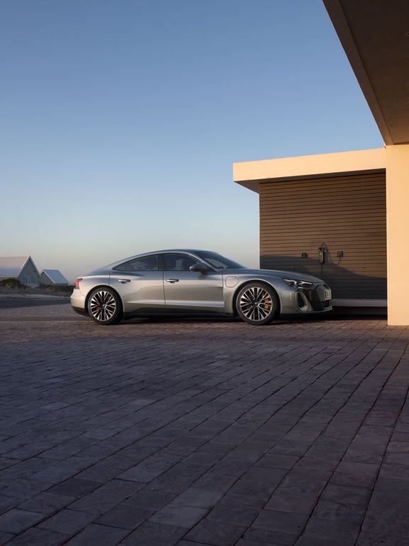 Ein silberner Audi S e-tron GT steht auf einer gepflasterten Auffahrt und ist an eine Ladestation angeschlossen. Im Hintergrund sind moderne Häuser und eine Dünenlandschaft zu sehen.