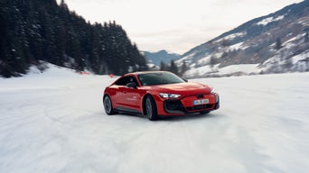 A red Audi RS e-tron GT performance speeds across a snow-covered area; snow dust, a forest, and mountains in the background.