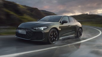 Dunkelgrüne Audi RS 5 Limousine fährt auf nasser Straße durch eine Kurve, LED-Scheinwerfer leuchten, Berglandschaft im Hintergrund.