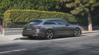 Grauer Audi A6 Avant parkt am Straßenrand vor einer Hecke und einem Baum bei Tageslicht in urbaner Umgebung.