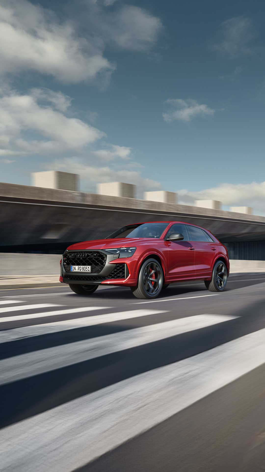 Ein roter Audi RS Q8 SUV performance fährt auf städtischer Straße mit Zebrastreifen, im Hintergrund modernes Gebäude mit Betonfassade.