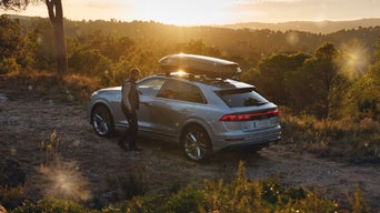 Audi Q8 mit Dachbox steht bei Sonnenuntergang auf Anhöhe, daneben ein Mann in der Natur.