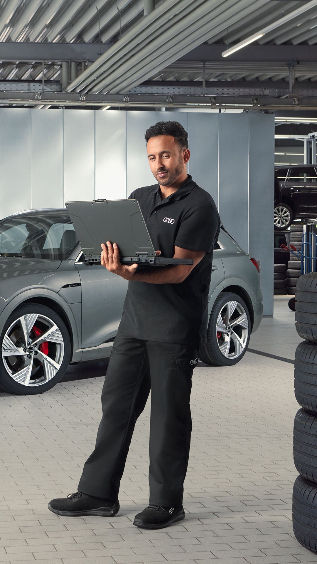 Audi-Mitarbeiter steht in Werkstatt vor einem grauen Audi Q4 e-tron und arbeitet an einem aufgeklappten Laptop.