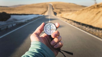 A hand holds a compass in front of a straight country road