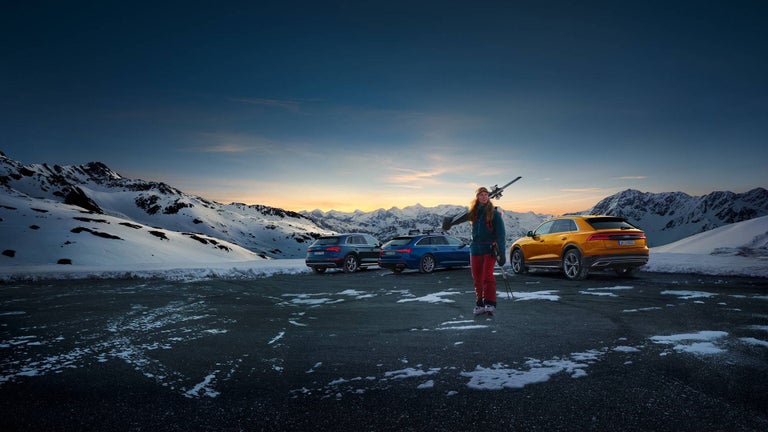A woman stands with skis in front of a mountainous landscape and various Audi models