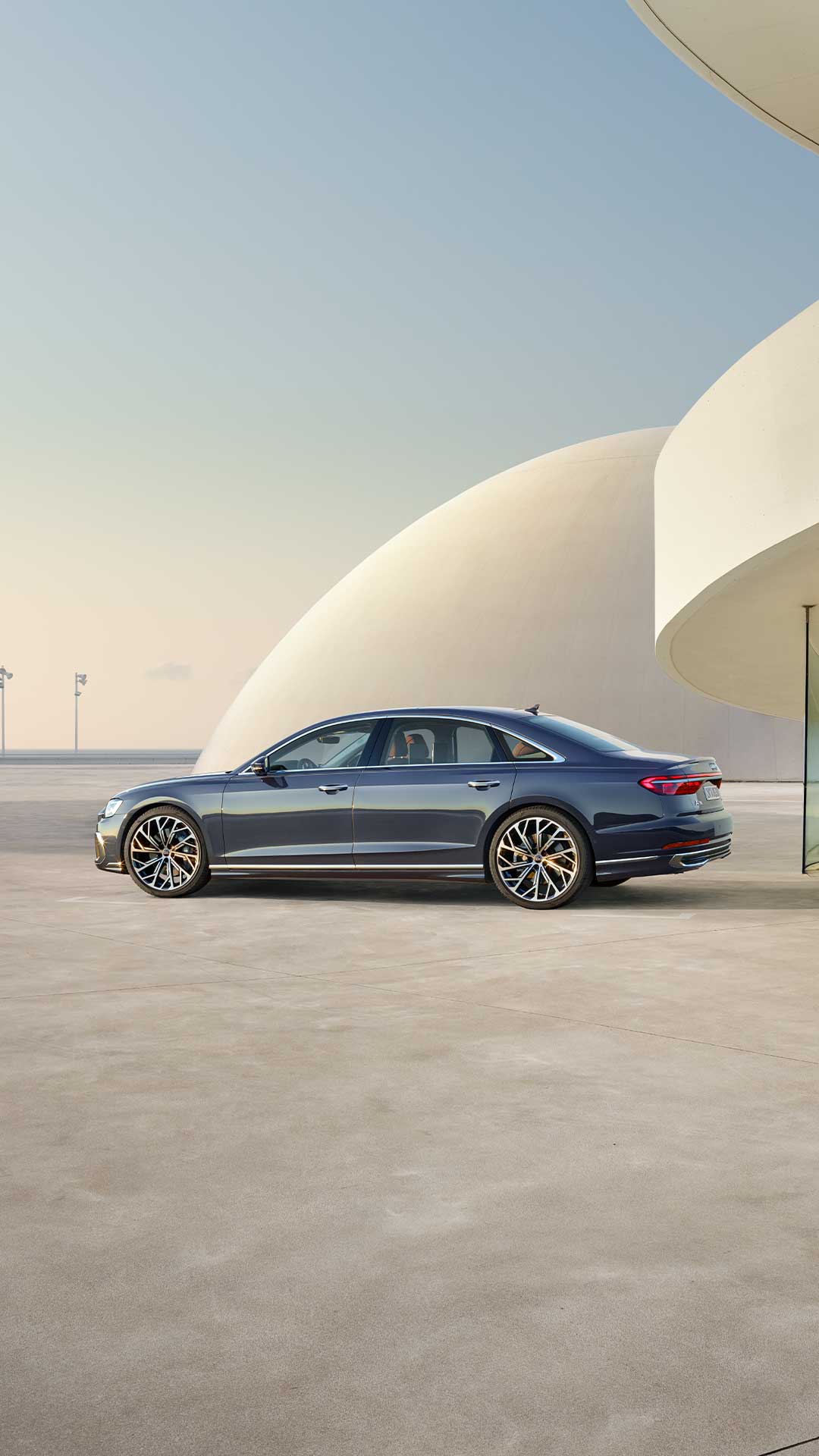 Ein dunkelblauer Audi A8 L steht seitlich vor moderner, heller Architektur bei Tageslicht.