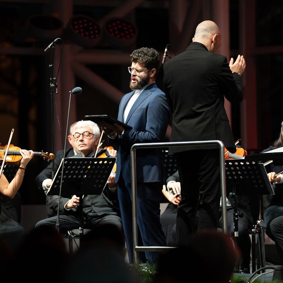 Solist liest aus einem Notenheft vor dem Orchester, neben ihm ein Dirigent; Musiker sitzen konzentriert unter warmem Bühnenlicht.