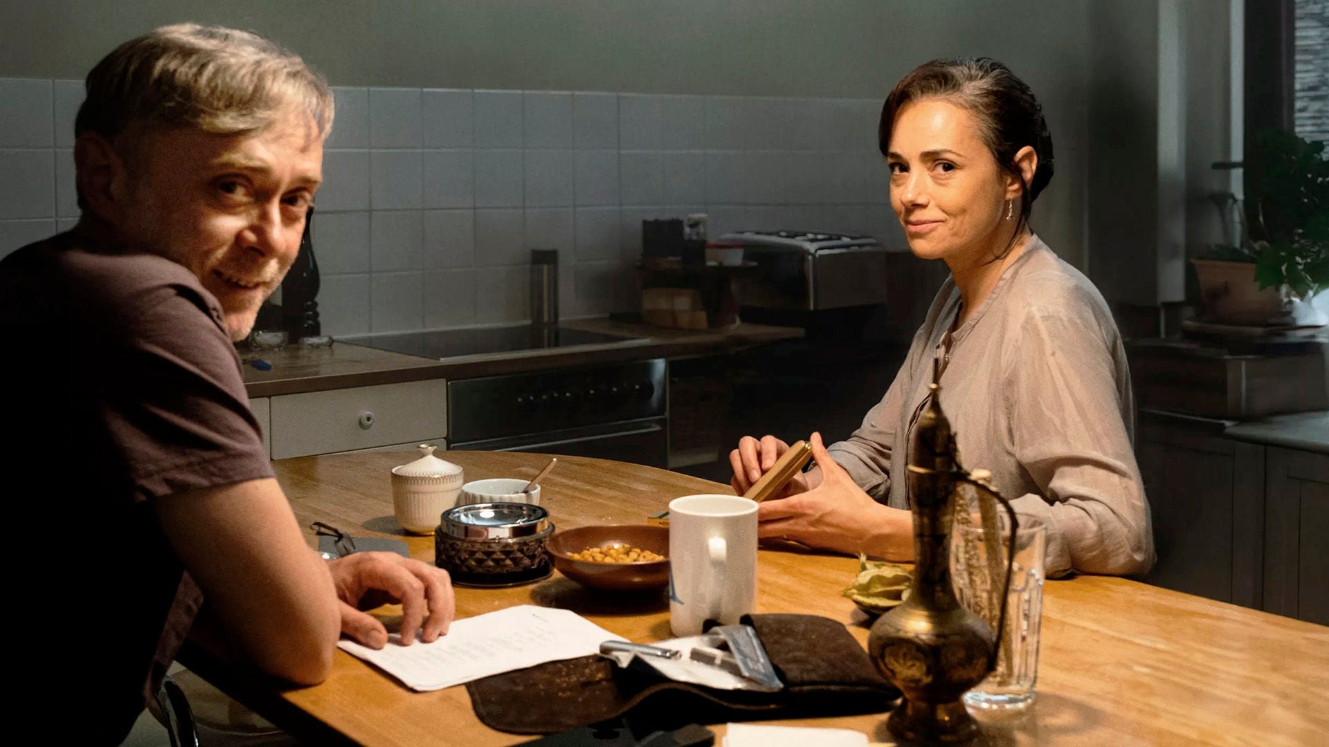 Ein Mann und eine Frau sitzen in der Küche an einem Tisch und lächeln den Zuschauer an.