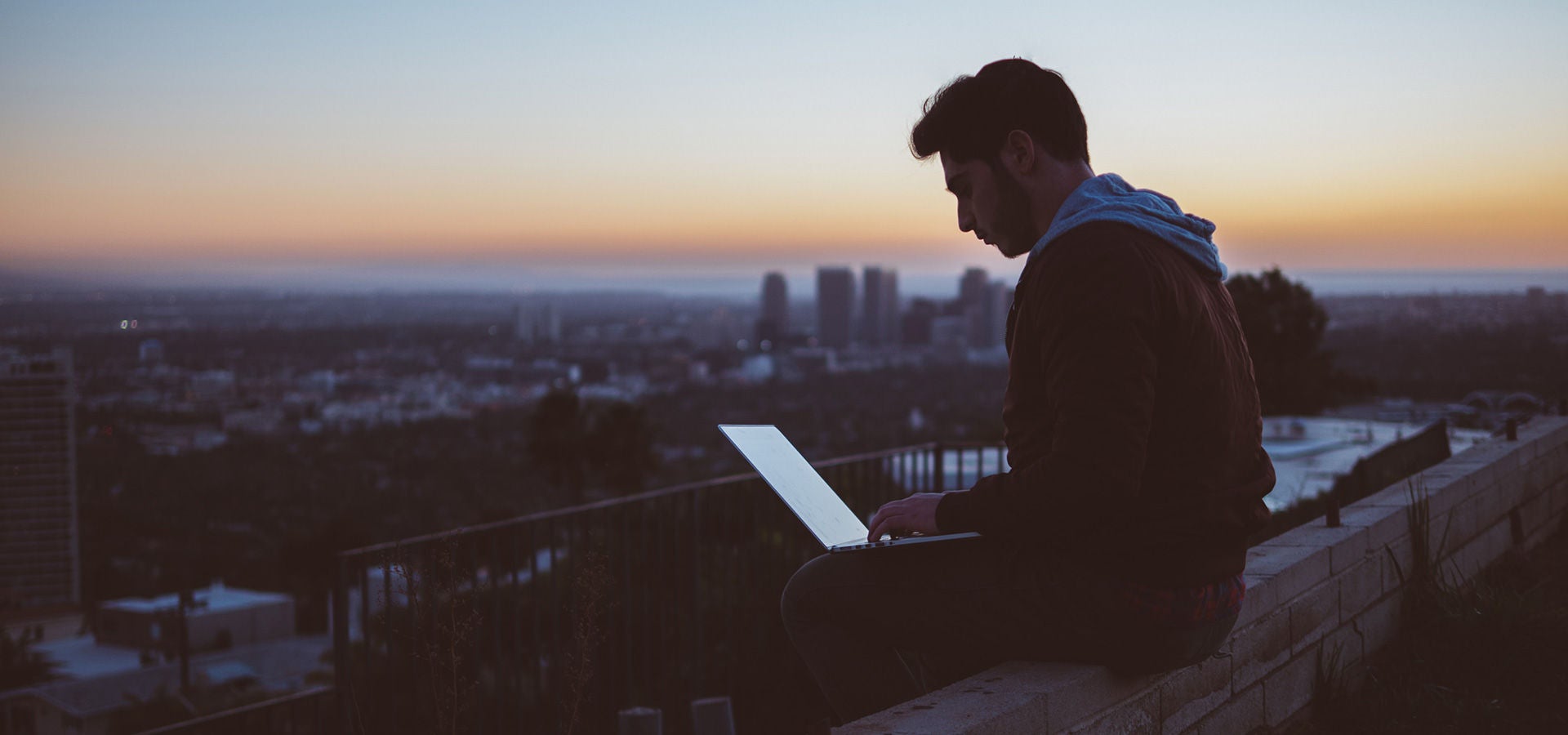Ein Mann sitzt bei Sonnenaufgang auf einer Mauer und hat seinen Laptop auf den Schoß