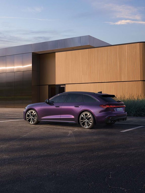 Lila Audi S5 Avant von der Seite, geparkt vor moderner Fassade mit Frau im Hintergrund.