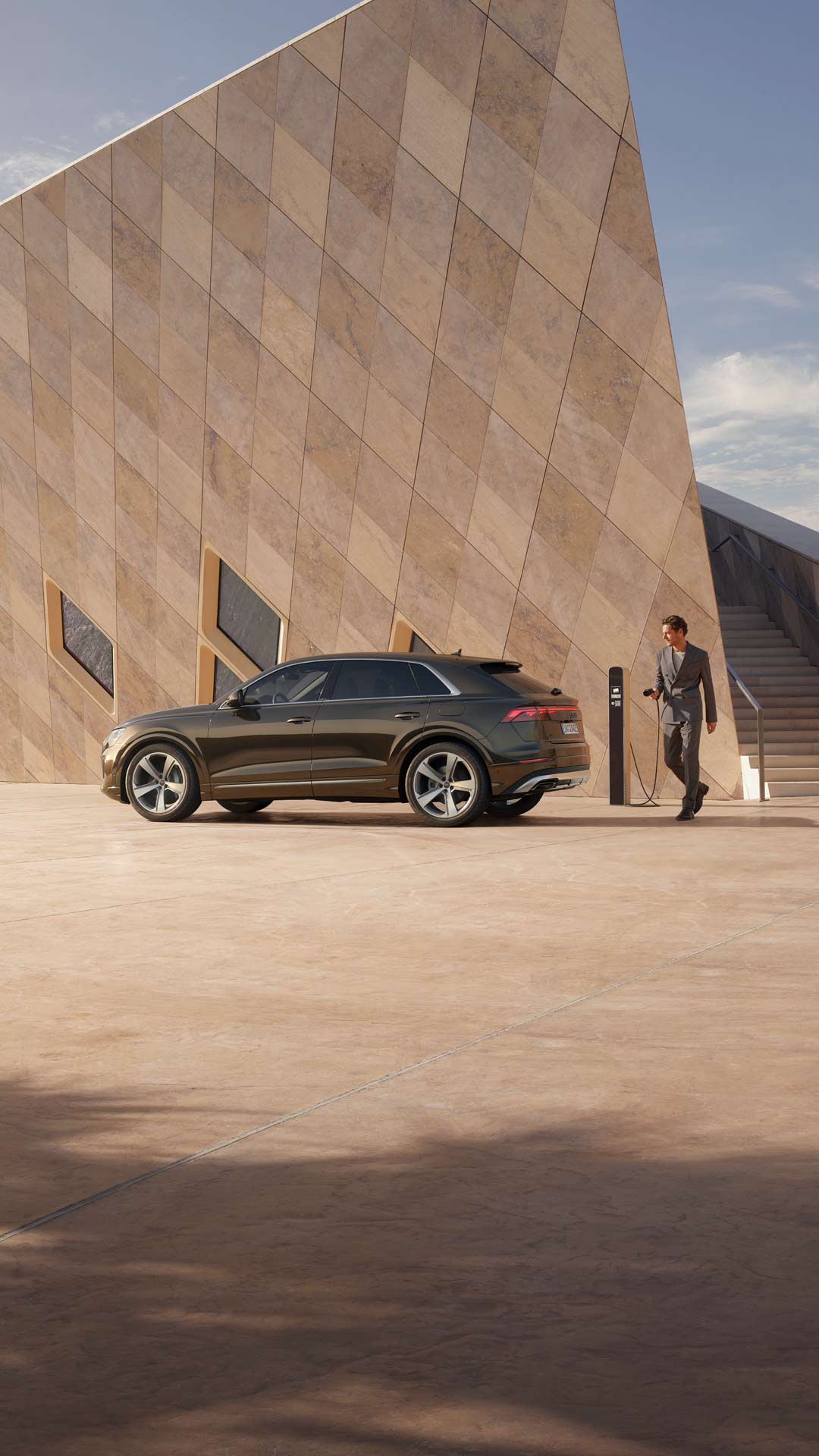 Ein Mann im Anzug geht auf eine Ladesäule zu, neben ihm ein brauner Audi Q8 SUV TFSI e vor moderner Architektur.
