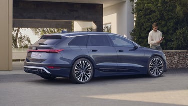 Der S6 Avant e-tron steht schräg seitlich auf Pflastersteinen vor einem modernen Gebäude. An einer am Gebäude befestigten Steinmauer hinter dem Fahrzeug ist ein Mann mittleren Alters gelehnt.