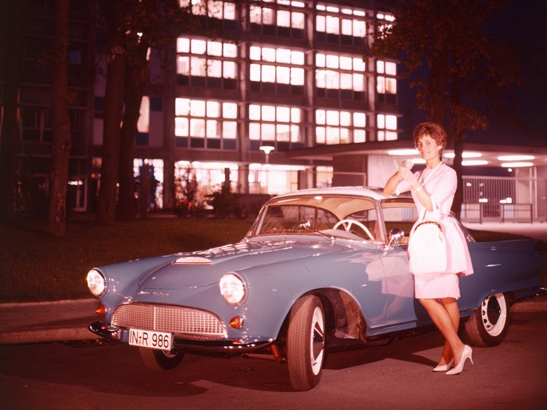A woman in a dress leans against a blue Union 1000 SP car in front of a house