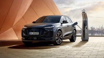 Seitliche Front des SQ6 SUV e-tron, welcher neben einer orangenen Gebäudefassade an eine Ladestation angeschlossen ist. Im Hintergrund ist das Meer, sowie die Skyline einer Stadt zu erkennen.