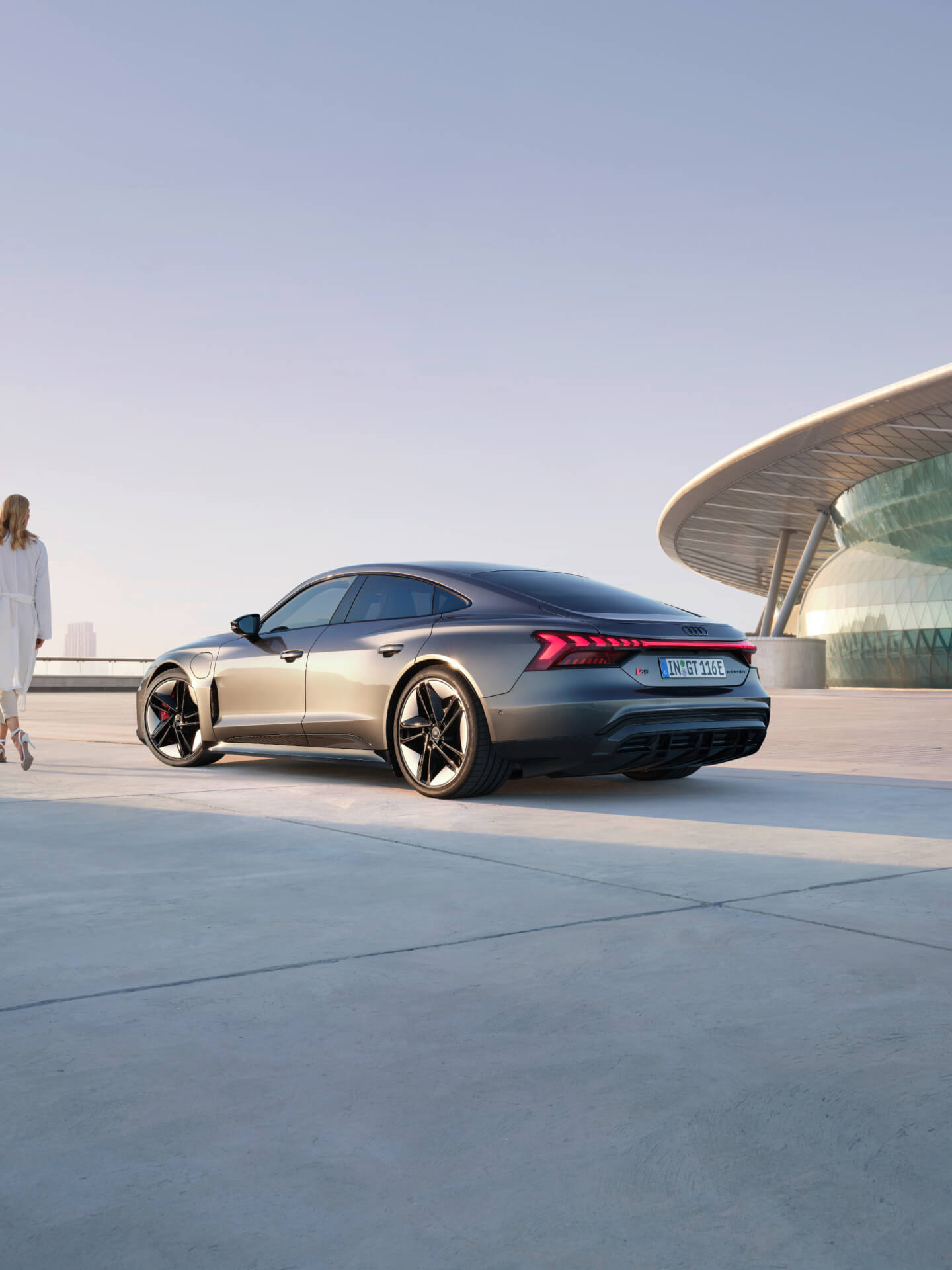 Dark grey Audi car being charged outdoors, by the sea, with a woman standing behind it