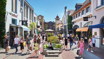 Viele Menschen schlendern durch eine sonnige Outlet-Shoppingmeile mit bunten Geschäften und Pflanzen.