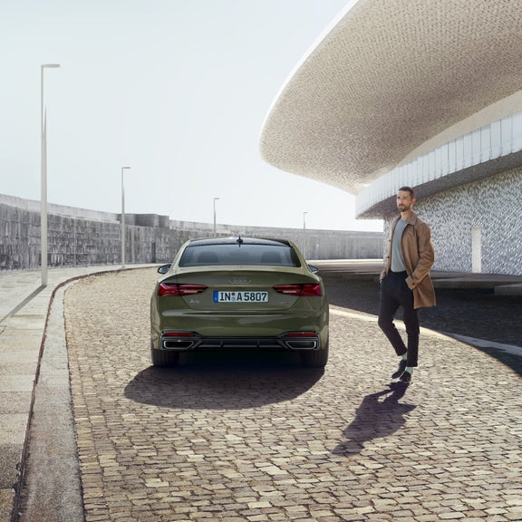 A green Audi car parked on a cobblestone road with a person standing beside it, under modern architecture.