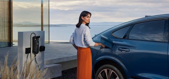 Frau lädt einen blauen Audi SQ6 Sportback e-tron an einer Wandladestation auf, im Hintergrund Meerblick und moderne Architektur.