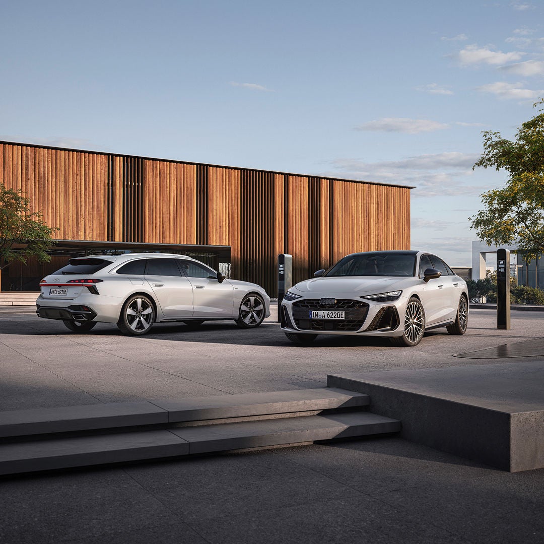 Two Audi cars in a stylish plaza with a wooden building and charging stations.