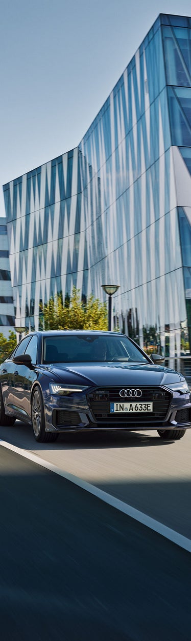 Schwarzer Audi A6 fährt auf einer städtischen Straße vor einem modernen Glasgebäude bei Tageslicht.