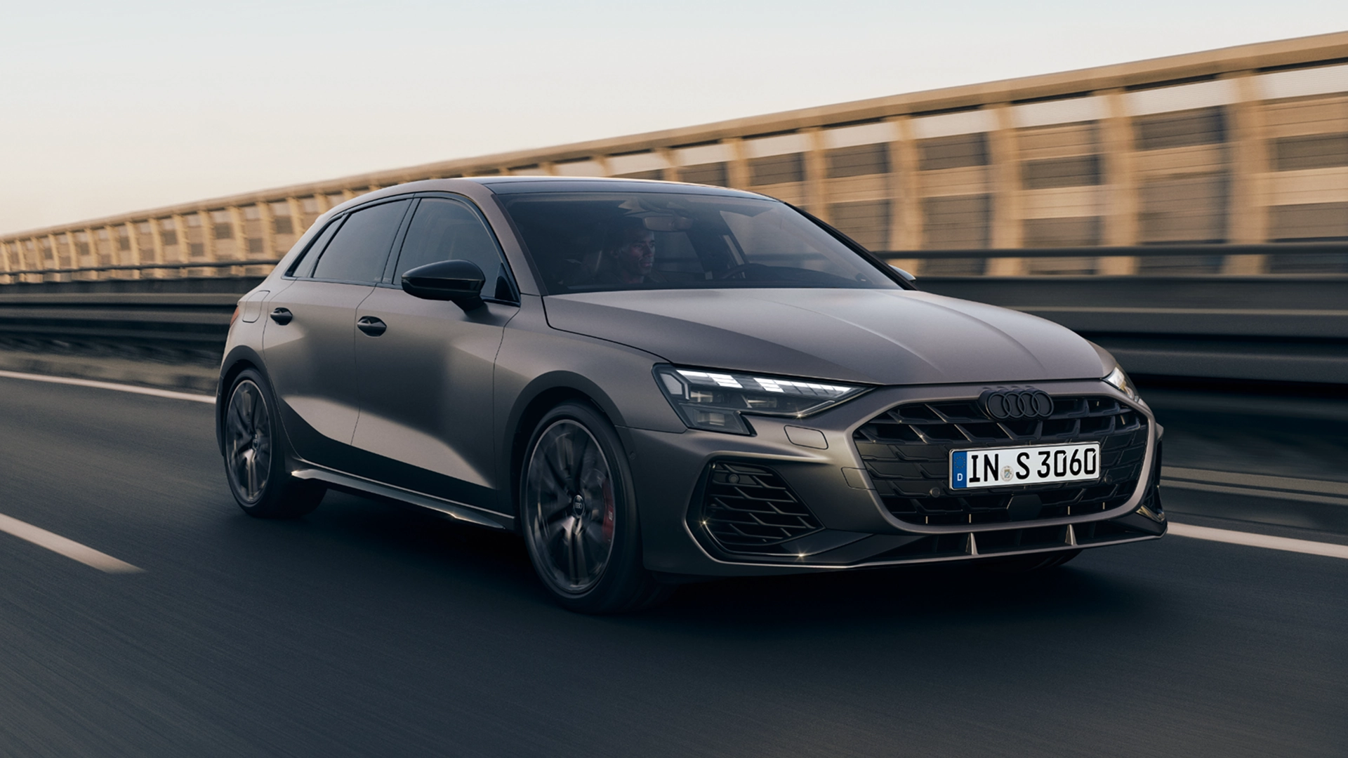 Grey Audi S3 Sportback driving on road, centre of image, with bridge and guardrail in background.