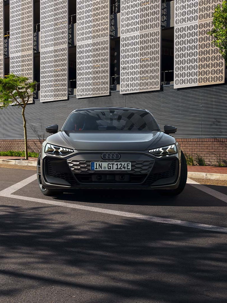 Ein grauer Audi RS e-tron GT steht frontal auf einer Straße mit moderner Metallfassade und Bäumen im Hintergrund.