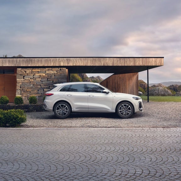 A white SUV parked beside a modern stone and wood house, set against a scenic backdrop of hills and clouds.