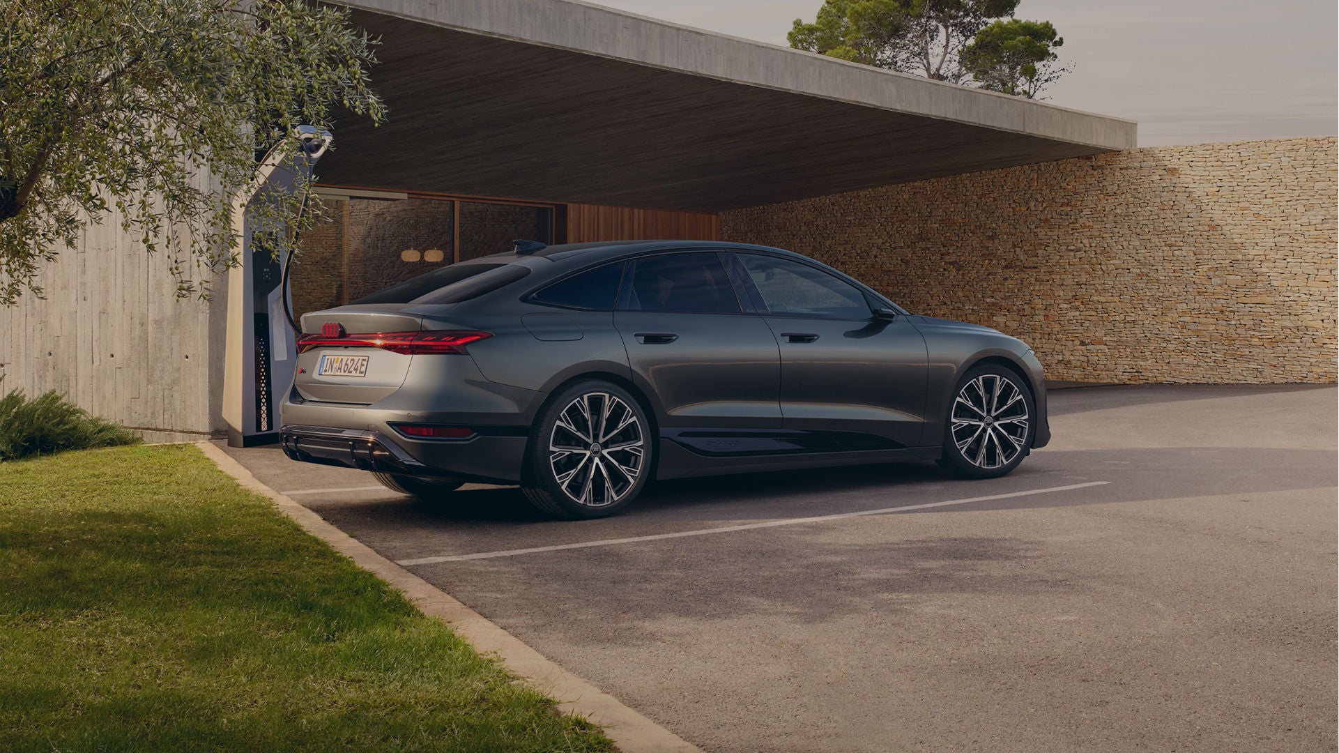 A sleek grey sedan parked outside a modern house under an open garage, near trees and a stone wall.