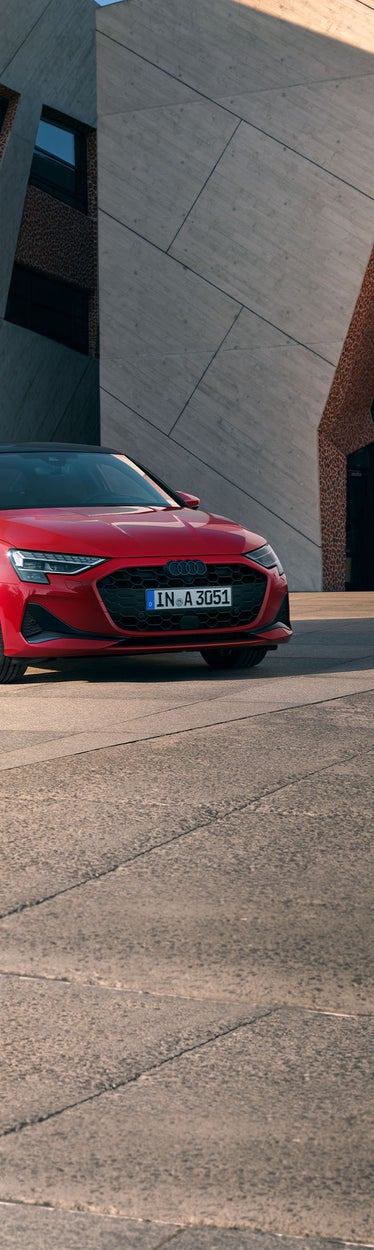 Eine rote Audi A3 Limousine steht auf einem hell gepflasterten Platz vor einer modernen Fassade mit Mosaik- und Betonstruktur.