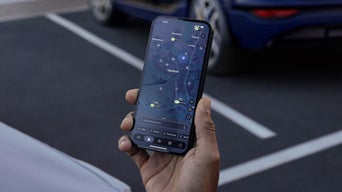 Hand hält Smartphone mit Ladepunktsuche, im Hintergrund das Heck eines Audi-Fahrzeugs.