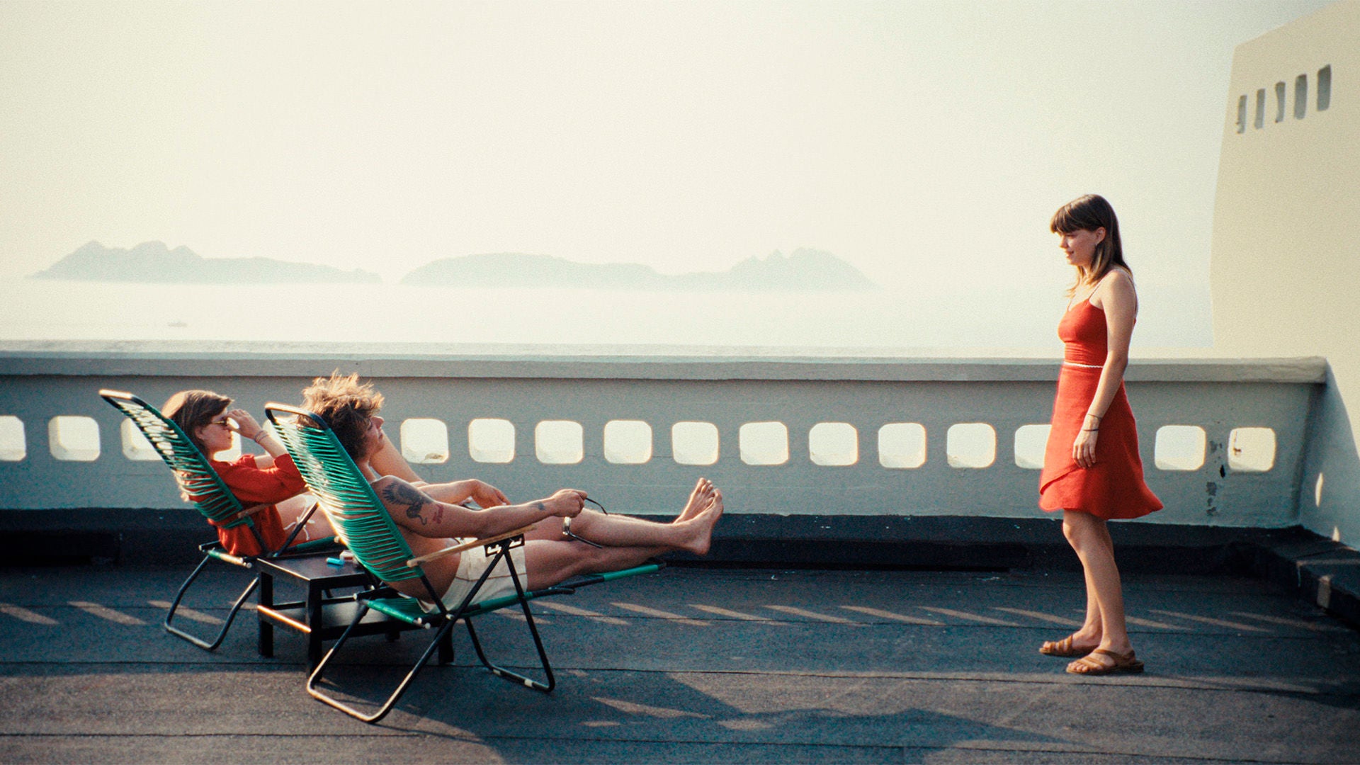 Drei Frauen auf dem Deck eines Schiffes: Zwei liegen nebeneinander auf Liegestühlen, die Dritte steht ihnen gegenüber.