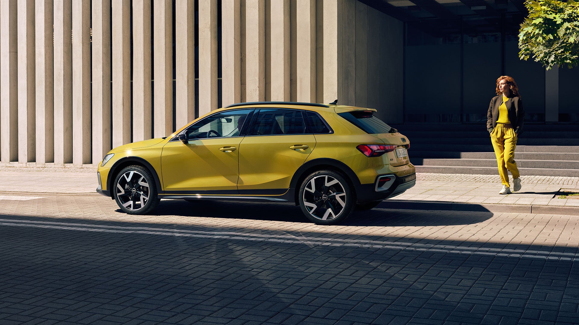 Gelber Audi A3 steht seitlich vor moderner Betonfassade, eine Frau im gelben Outfit geht auf den Wagen zu.         