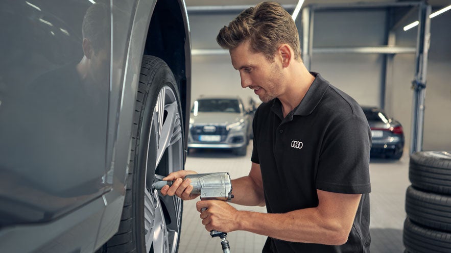 Mechaniker in Audi-Arbeitskleidung zieht mit einem Schlagschrauber eine Radmutter an einem Fahrzeug in der Werkstatt fest.