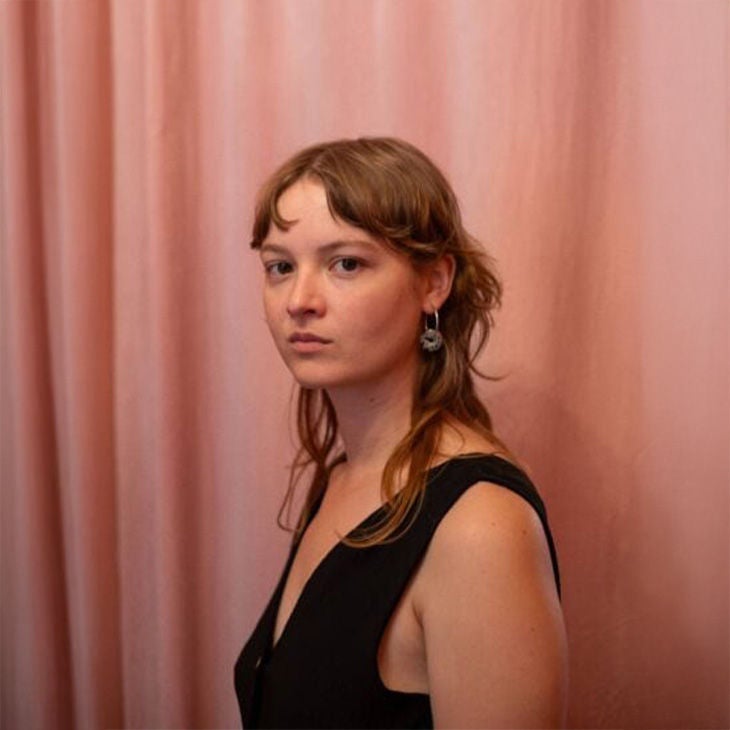 Frau mit heller Haut und dunklem Top steht vor rosa Vorhang und blickt ruhig in die Kamera.