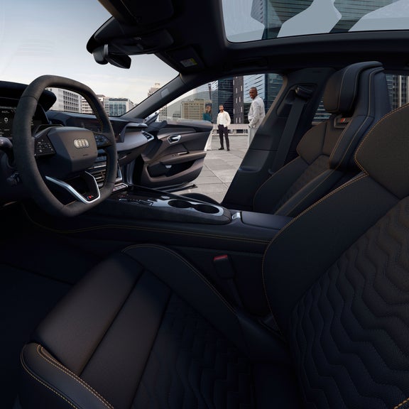 Innenraum des Audi S e-tron GT mit sportlichem Lenkrad, digitalen Anzeigen und hochwertigen Materialien. Durch das offene Fenster sind drei Personen auf einer Dachterrasse mit Stadtblick zu sehen.