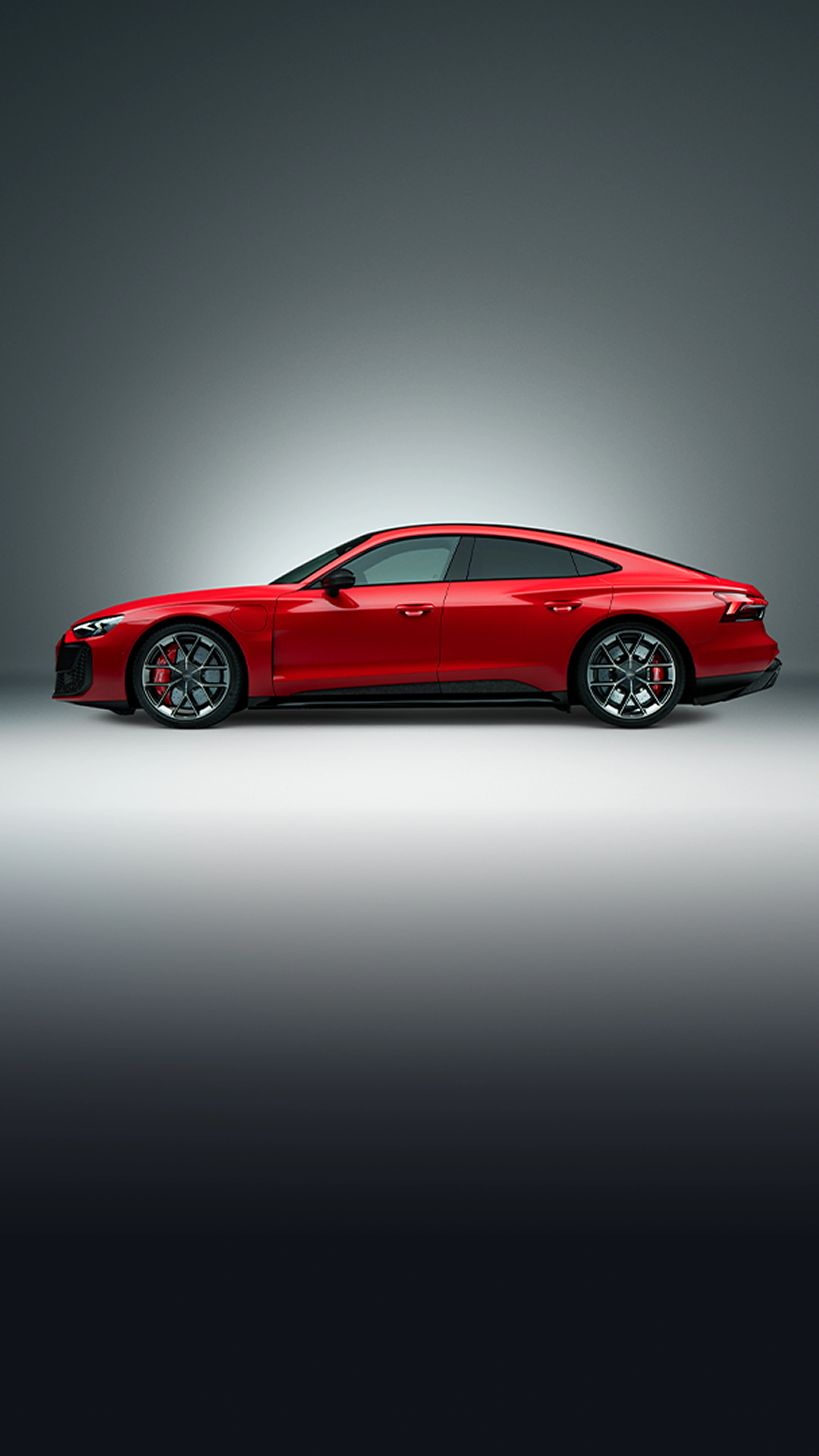 Red Audi e-tron GT Sportback shown in full side profile against a neutral grey studio background. The vehicle features a low, aerodynamic fastback silhouette, frameless windows, large multi-spoke alloy wheels, red brake callipers and sculpted body lines. Studio lighting highlights the sporty proportions and premium design details.