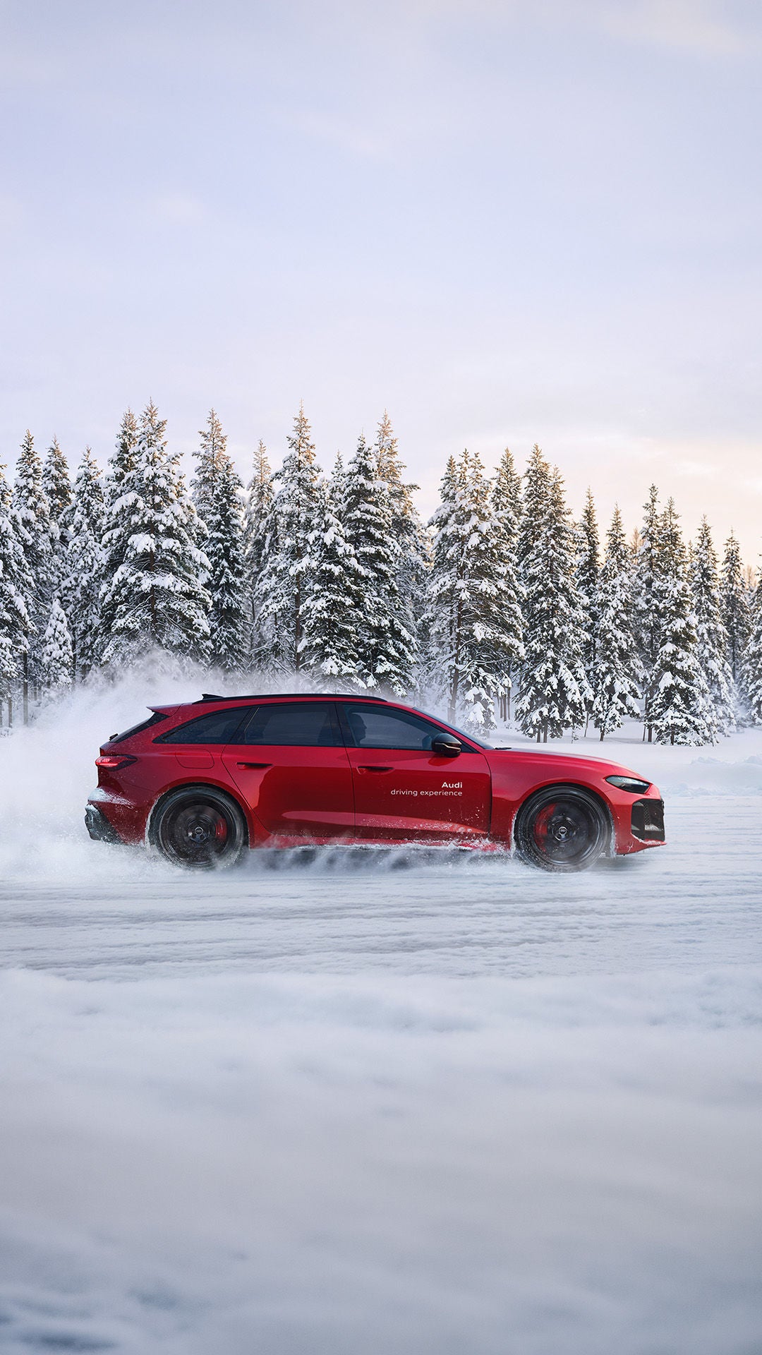 A red Audi RS 5 Avant driving on snow; snow dust, snow-covered fir trees, and a bright sky in the background.