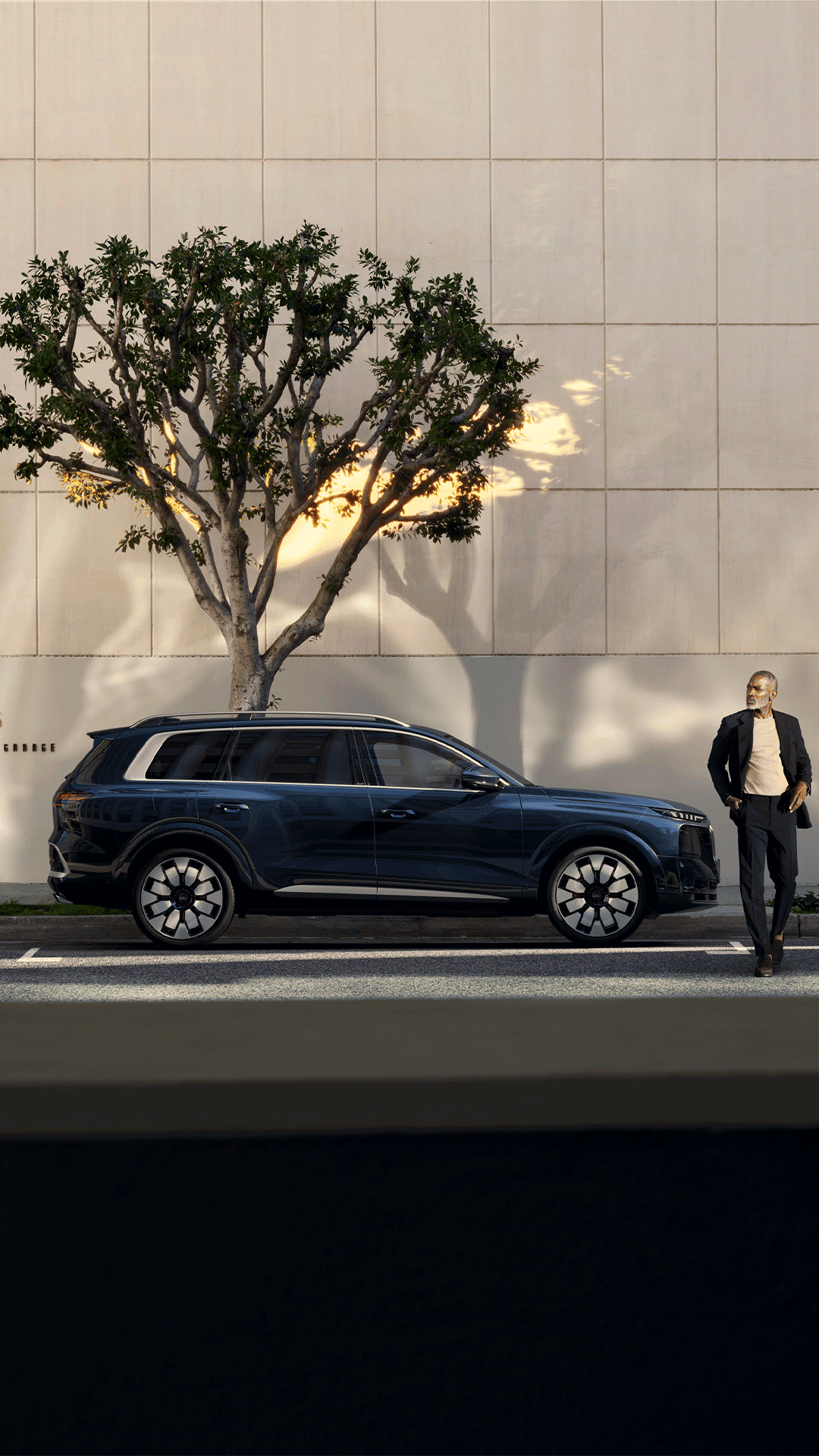 Q9 SUV bleu foncé est stationné devant un bâtiment moderne aux murs clairs. Un arbre projette des ombres sur la façade, tandis qu’une personne se tient à proximité du véhicule sur le trottoir. La scène est éclairée par la lumière du jour dans un environnement urbain contemporain.