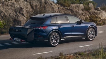 Blauer Audi Q4 e-tron fährt mit großem schwarzen Heckgepäckträger auf Landstraße entlang felsiger Landschaft.