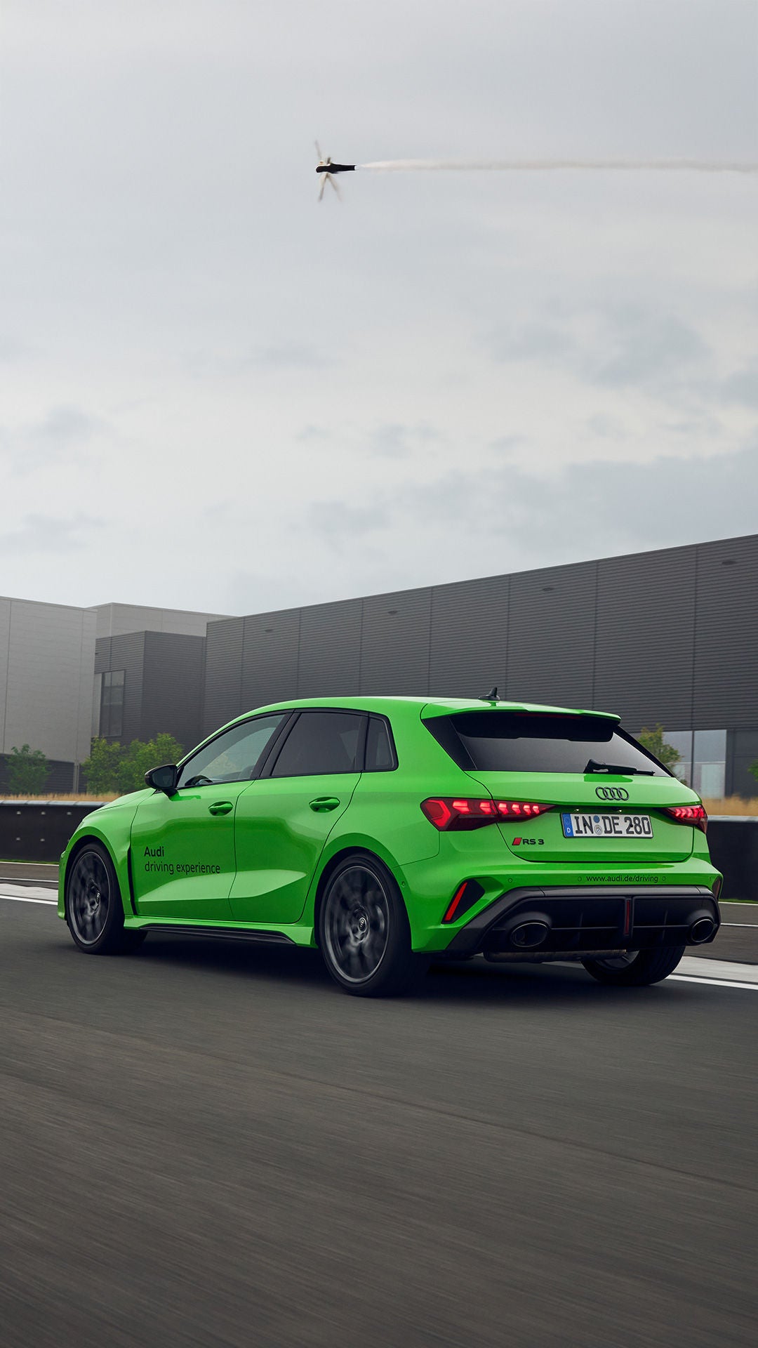 Hellgrüner Audi RS 3 Sportback fährt auf einer Teststrecke in Neuburg, im Hintergrund ist ein modernes Gebäude mit Audi Sport Logo.