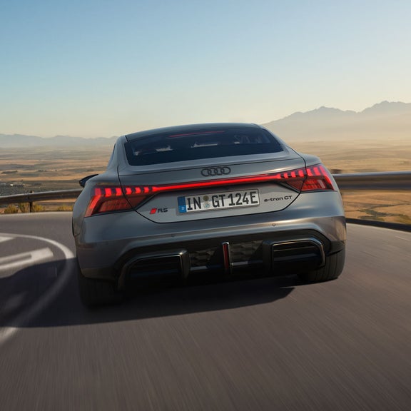 Ein grauer Audi RS e-tron GT frontal von hinten durchquert die steile Kurve einer Landstraße. Im Hintergrund eine ländliche Umgebung und Gebirgszüge.