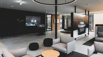 Heller Loungebereich mit grauen Sesseln, runden Tischen und großen Bildschirmen mit Audi-Bildmotiven an den Wänden.