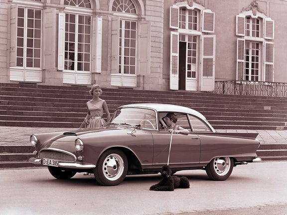 Eine junge Person sitzt am Steuer eines Auto Union 1000 SP und hat einen Pudel an der Leine, der vor dem Auto liegt. Eine Frau steht neben dem Wagen.