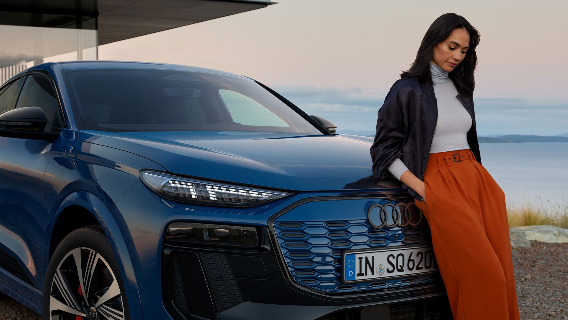 Dunkelhaarige Frau lehnt am blauen Audi SQ6 Sportback e-tron vor moderner Architektur, im Hintergrund Wasser und Horizont.