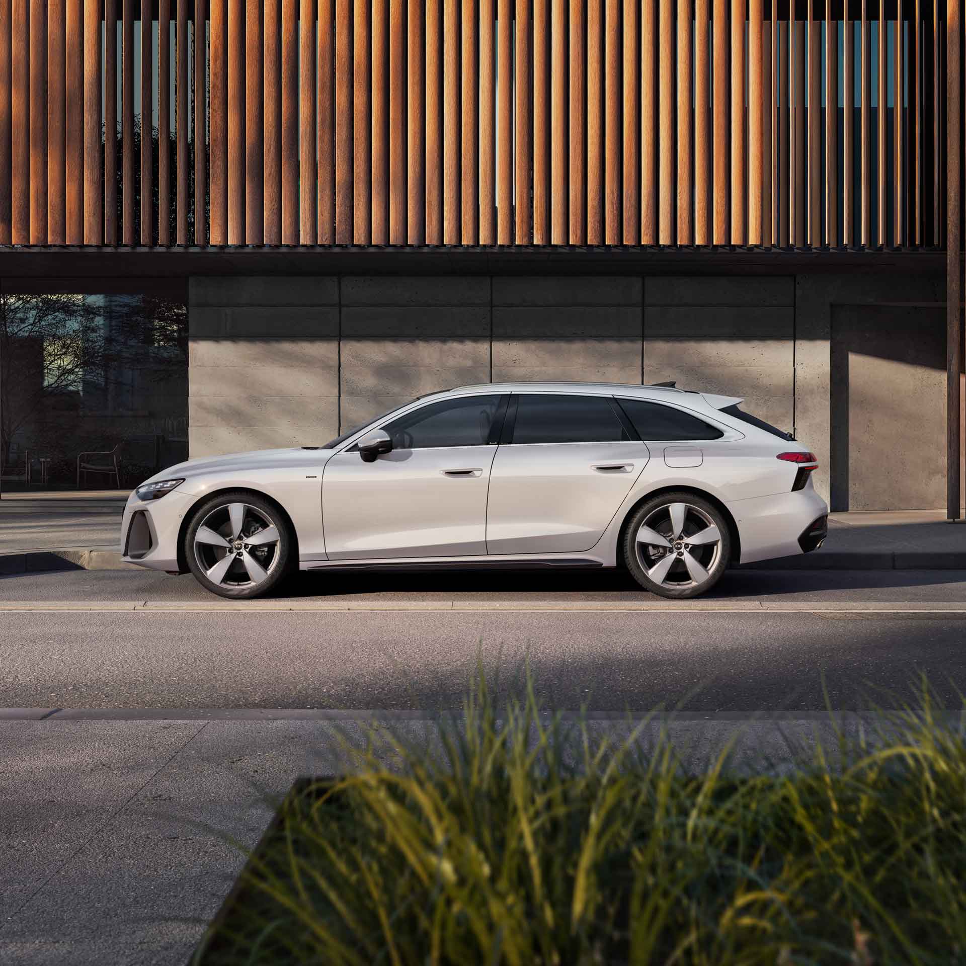 Vit Audi A6 Avant e-hybrid sedd från sidan på en parkering. En byggnad med träpaneler i bakgrunden.