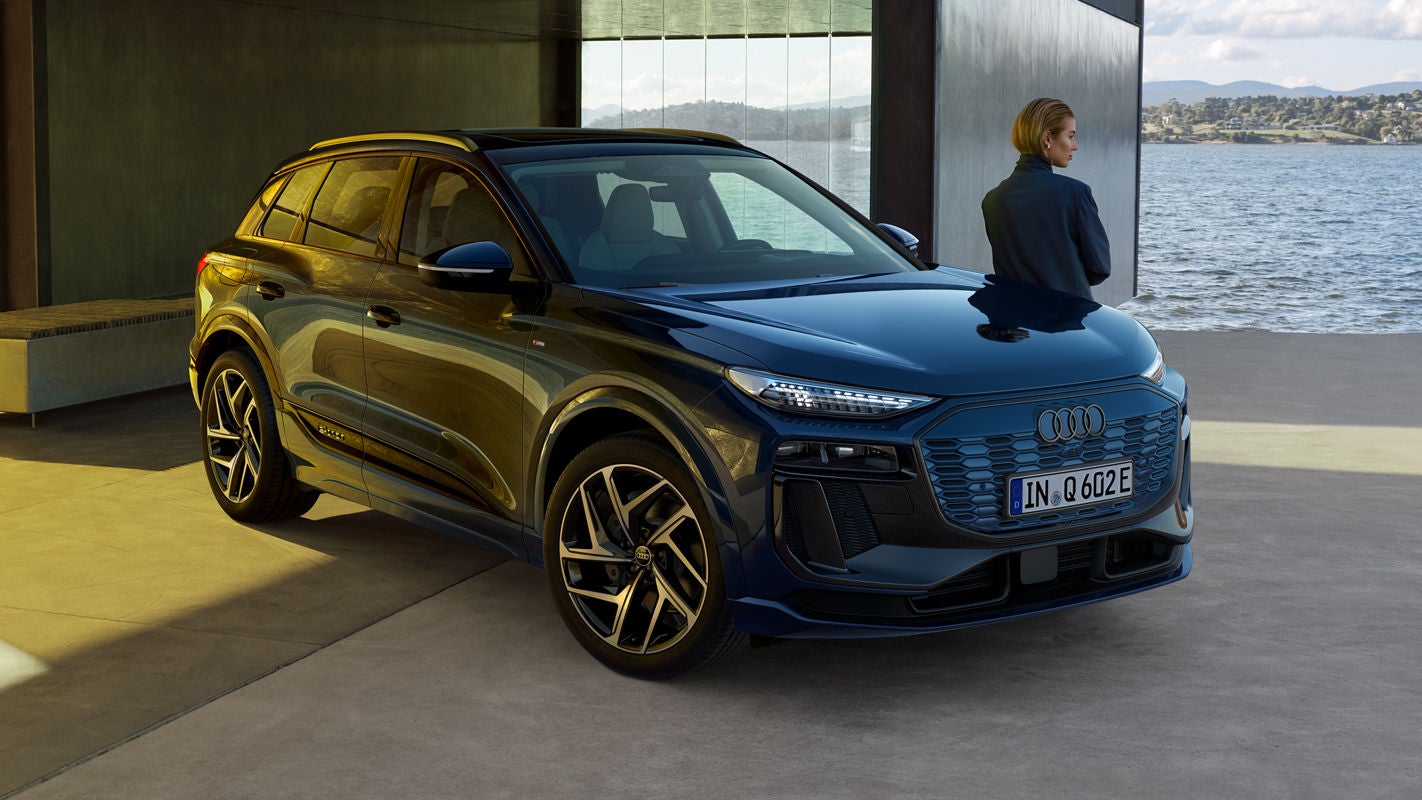 Ein blauer Audi Q6 SUV e-tron steht vor einem Gebäude am Wasser, eine Frau, mit kinnlangem Haar, blickt über die Landschaft.